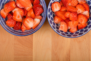 Two patterned bowls filled with sliced fresh strawberries are positioned side-by-side on a light...