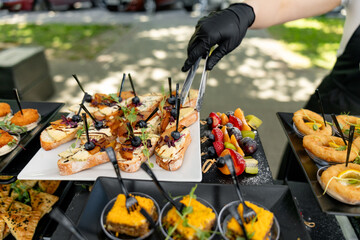  Cook puts snacks on table. Catering. Festive buffet. Table setting. Desserts and snacks on catering table. Waiter in black apron with tray. Bruschetta, desserts with fruit in baskets. Front view