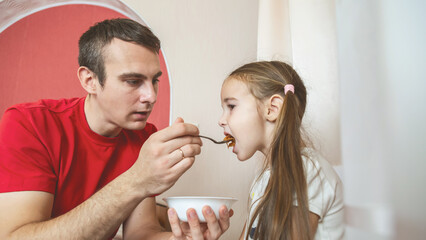 Cheerful small girl daughter and her handsome young dad spending time together at home. Father feeding his little daughter.
