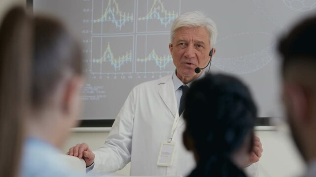 Medium shot from audience space of senior male clinical professor equipped with earset microphone delivering lecture in auditorium sharing latest developments with colleagues during group seminar