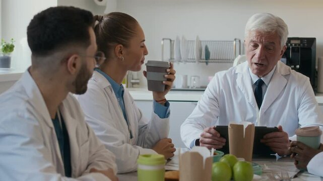 Medium shot of senior male chief physician engaging in lively discussion with clinical staff team during coffee break in hospital kitchen