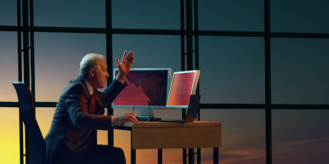Businessman sitting at desk, raising hand in frustration while looking at two computer screens...