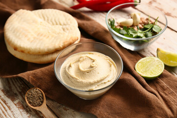 Bowl of tasty hummus with ingredients, bread and napkin on light wooden background, closeup