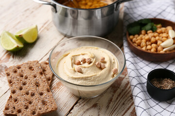Bowl of tasty hummus with ingredients, crackers and napkin on light wooden background, closeup