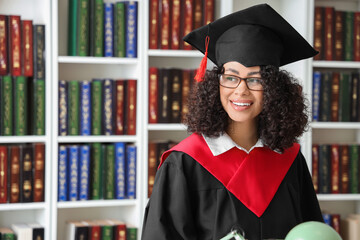 Pretty African-American female graduate in eyeglasses near bookshelves in library