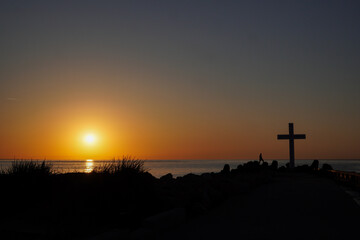 Obraz premium A serene sunset casts vibrant hues over the ocean while a lone figure stands next to a prominent cross on the shore, surrounded by rocky formations and grass.
