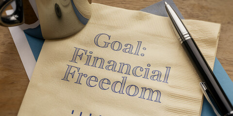 Beige napkin with blue text reading "Goal: Financial Freedom", accompanied by a pen and cup reflection, suggesting ambition and financial planning