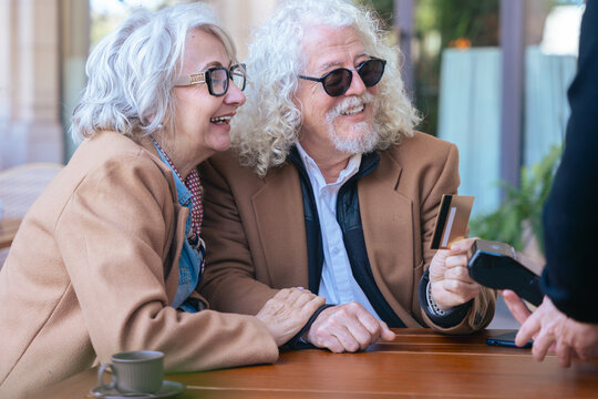 Smiling senior couple makes a credit card payment at an outdoor cafe. They wear stylish coats and sunglasses, enjoying a relaxed moment together in a sunny, casual and urban environment. 