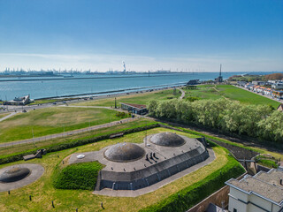 Obraz premium Aerial view of historic coastal fort with bunkers, grassy landscape and museum buildings against wide river, canal, bay with industrial port and cranes in background under clear blue sky.