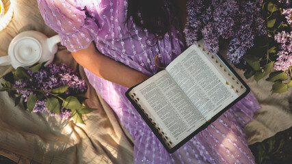 Woman in a lilac dress reading the Bible outdoors, surrounded by blooming lilacs, a white teapot,...