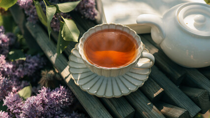 Vintage teacup filled with tea on a wooden bench surrounded by lilac flowers and a white teapot....