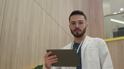 Medium portrait shot of young male healthcare expert of mixed ethnicity looking at digital tablet screen then turning to camera captured in hospital setting, slow motion