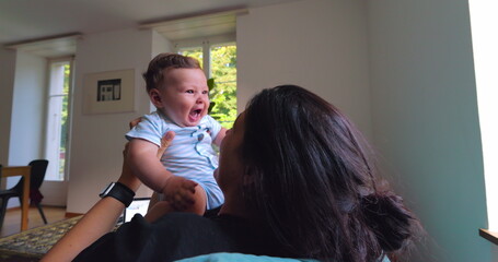 Mother lifting baby into the air while playing, smiling and laughing in a bright living room filled...