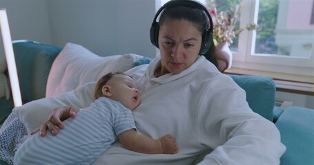 Mother multitasking with baby sleeping on her chest, wearing headphones, working on laptop, cozy...
