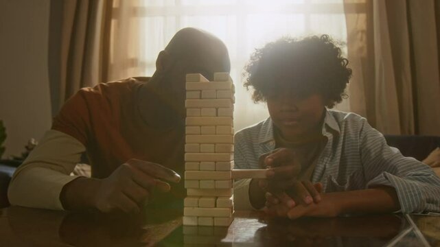 Joyful African American father and son focusing on stacking block game, sharing laughter and teamwork in their cozy living room