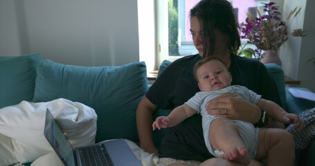 Obraz premium Mother working from home on a laptop with baby on her lap, enjoying a calm moment together on the sofa, with natural light filling the cozy living room