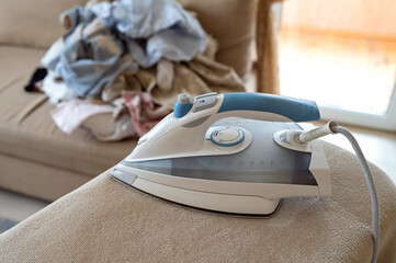 Modern steam iron ready to use, background shows pile of un-ironed laundry in living room