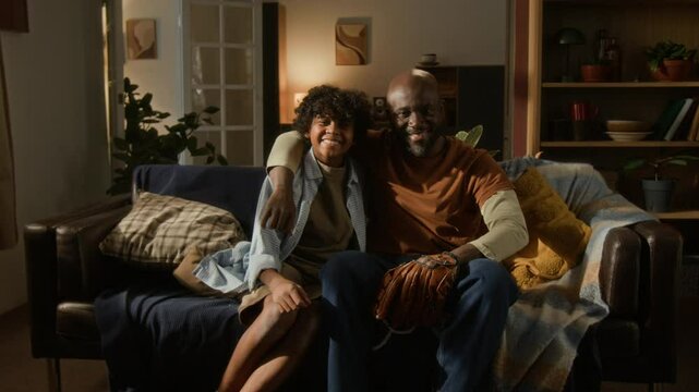 After quick game of catch, smiling African American boy and his father sitting close on couch, dad still holding baseball glove, both posing for camera with smile in their cozy living room