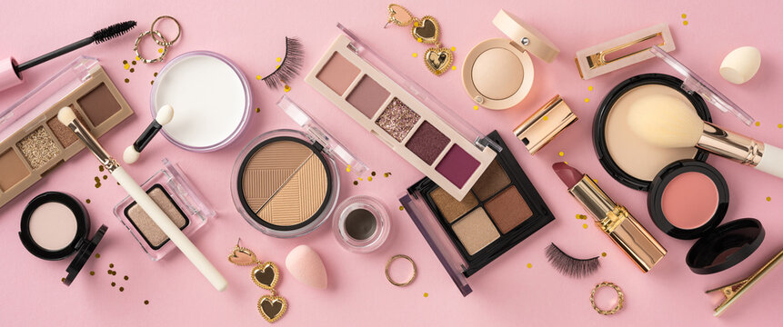 Flat lay of makeup essentials including palettes, brushes, and accessories on a stylized pink surface