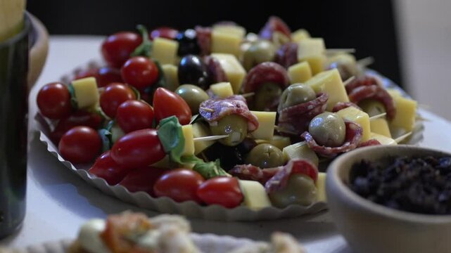 Argentine charcuterie board featuring basil‑topped cherry tomato skewers, cubed cheese and green and black olives wrapped in salami slices, arranged on a white platter in rustic setting, slow motion