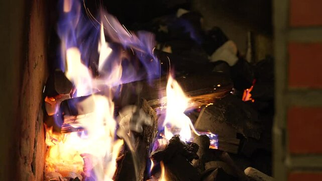 Seasoned firewood logs ignite in brick-lined firebox as yellow blue flames swirl around charred wood and glowing embers, casting warm flickering light on the rough masonry and blackened coals beneath