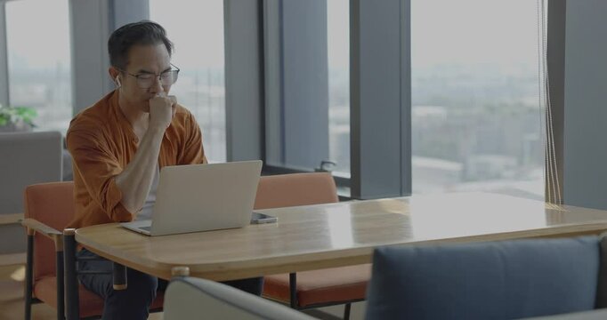 Senior Asian businessman wearing Bluetooth headset celebrates success during online meeting in modern co-working space. Remote work and business communication concept.