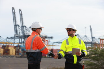 Two men with green safety vests and PPE are working at the port, using the logistics export-import...
