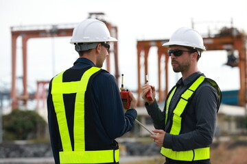 Two men with green safety vests and PPE are working at the port, using the logistics export-import...