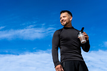 Fit man wearing black sportswear holding protein shake smiling confidently under bright blue sky...
