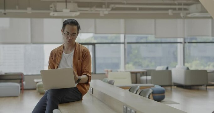 Senior Asian businessman wearing Bluetooth headset celebrates success during online meeting in modern co-working space. Remote work and business communication concept.