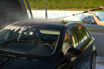 A person is washing a black car with a high-pressure water hose, focusing on the roof. Soap suds are visible on the car, and the car is parked outdoors during cleaning.