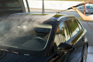 Obraz premium A person is using a high-pressure water hose to wash a black car outdoors. Water sprays across the windshield, creating a misty effect while ensuring the vehicle gets a thorough clean.
