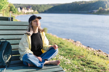 Woman meditating in sunny nature, relaxing, sitting barefoot on bench by river in butter yellow...