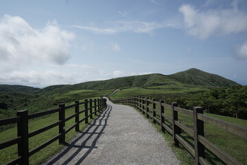 Obraz premium Sunlit gravel path, lined by wooden fences, winds through rolling green hills to a distant peak. Evokes peace, adventure, and the beauty of the open countryside. 