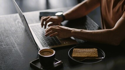 Close-up image of a person working on laptop computer at a table, typing on the laptop keyboard, with a cup of coffee and cake on the plate.