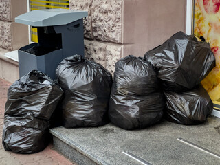 Black garbage bags are stacked next to a public trash bin outside a shop on a busy city sidewalk...