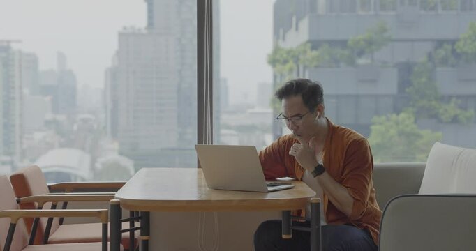 Senior Asian businessman wearing Bluetooth headset celebrates success during online meeting in modern co-working space. Remote work and business communication concept.