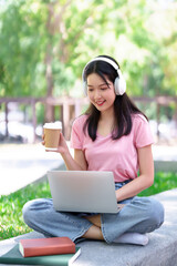 Young Asian Woman Studying Online Outdoors with Laptop Coffee and Headphones Enjoying Learning on...