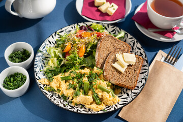 Scrambled eggs with vegetables, asparagus, toast, chives, seed sprouts and greens served on a beautifully decorated plate, a breakfast set, illuminated by the morning sun.