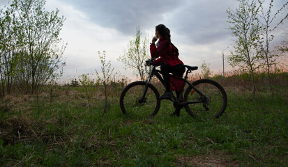 Young woman riding a bicycle on the spring field with green grass on sunset sky landscape background. Happy activity sport trip vacations
