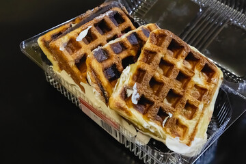 Sweet Belgian waffles in a plastic container on a black background (close-up). Baking of Belgian waffles and pastries.
