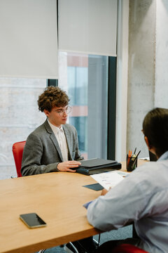 A young White man with glasses is having a discussion with a brunette woman during an internship interview in a modern office, sitting at a wooden table with documents and stationery.