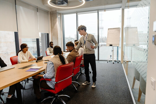 A young White man with glasses is leading a team discussion during an internship session with a multinational group of people in a modern office setting with a large wooden table and large windows.