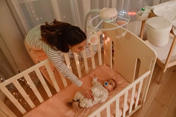 Fotobehang A white adult woman stands beside a crib, gently caring for her infant baby at home in a cozy nursery with soft lighting and a pacifier. © Drobot Dean