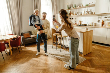 A white adult woman takes a photo of a man holding their son with a school backpack in a cozy modern kitchen. The family prepares for the school day during a cheerful morning.