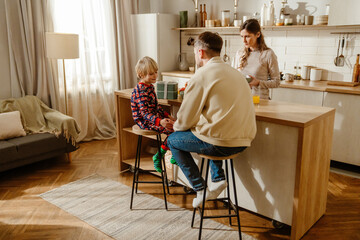Fototapeta premium A White man and woman are in a cozy, modern kitchen with their young son during breakfast. The child sits on a stool while the parents chat and prepare drinks at the island counter.