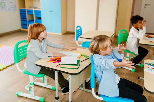 Three diverse school-aged boys are seated at desks in a bright classroom, discussing and gesturing expressively. Art supplies and notebooks are on their desks.