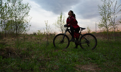 Fototapeta premium Young woman riding a bicycle on the spring field with green grass on sunset sky landscape background. Happy activity sport trip vacations