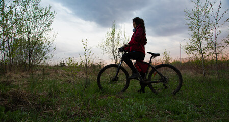 Young woman riding a bicycle on the spring field with green grass on sunset sky landscape...