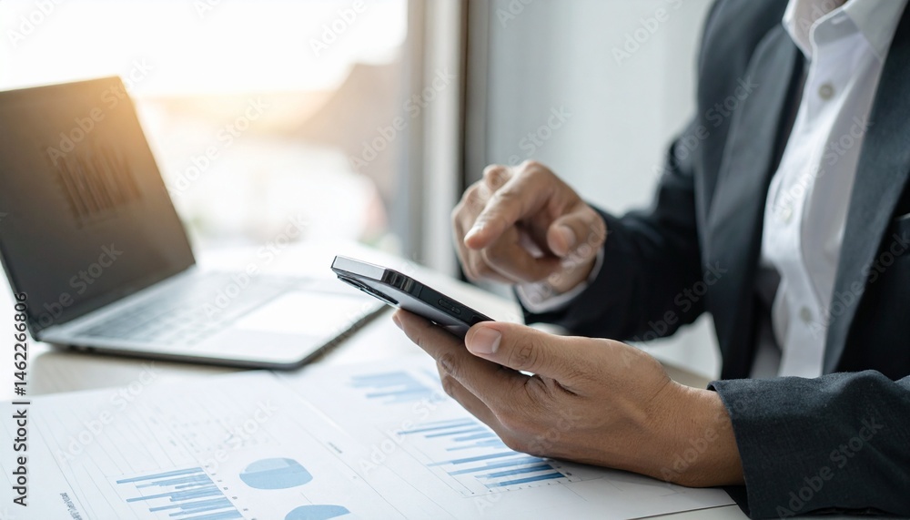 Poster a person in a suit using a smartphone while analyzing data charts on a desk, with a laptop in the ba